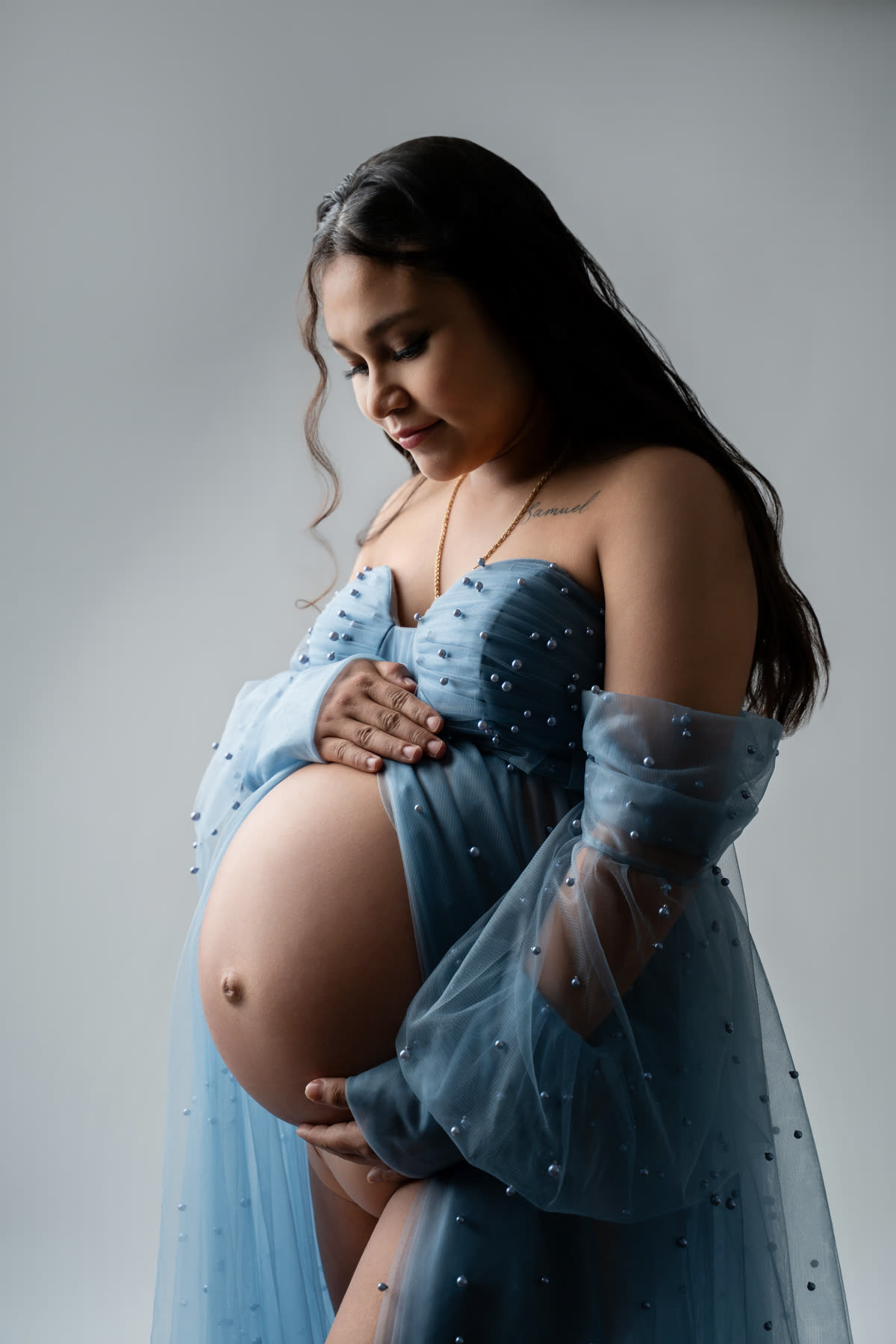 Expectant mother in a sheer blue gown looking down at her belly