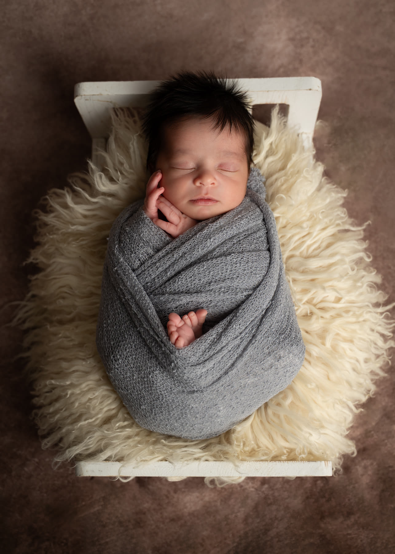 Newborn wrapped in gray in a tiny bed with fur